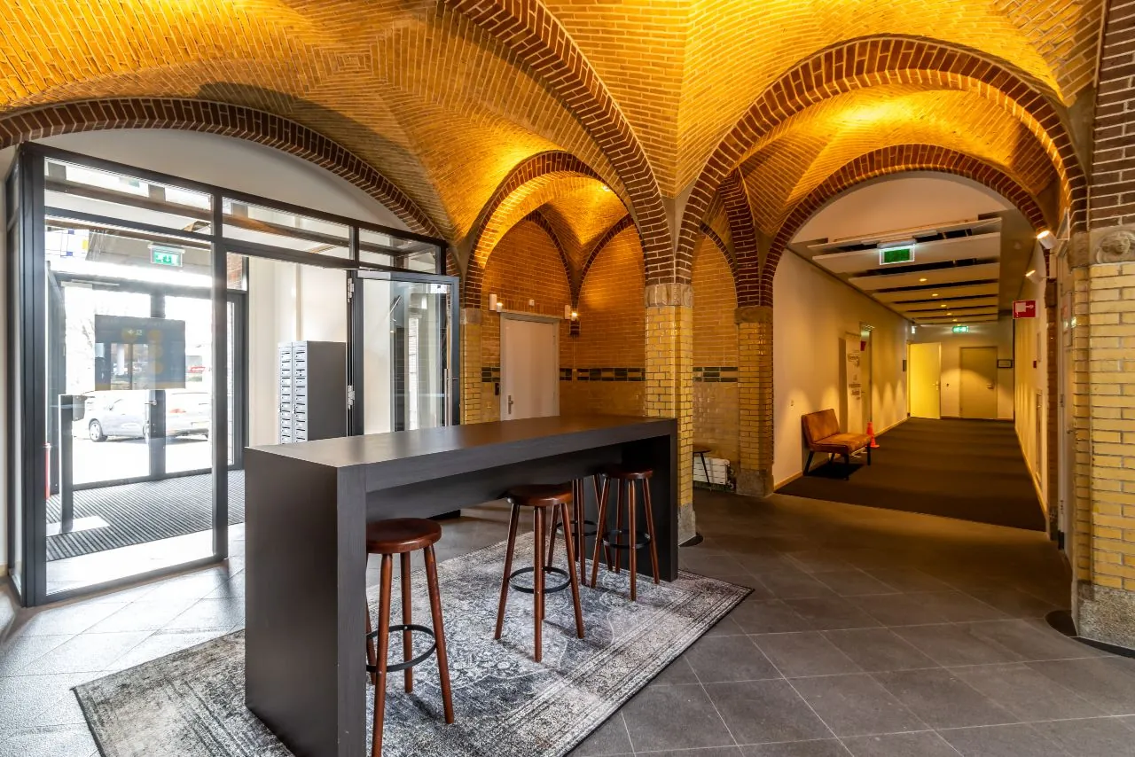 Entreehal van een kantoorpand aan de Lage Naarderweg met gewelfde bakstenen plafonds, een hoge tafel met krukken en een gang naar kantoorruimtes.